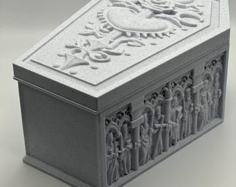 May include: A gray, coffin-shaped box with detailed relief carvings. The lid features a heart and rose design, while the sides depict figures within arched frames. The box has a textured surface and a gothic aesthetic.