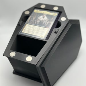 May include: Black coffin-shaped card game deck box with a magnetic closure. The box has a compartment for holding cards and a separate compartment for dice or tokens. The box is made of 3D printed plastic.