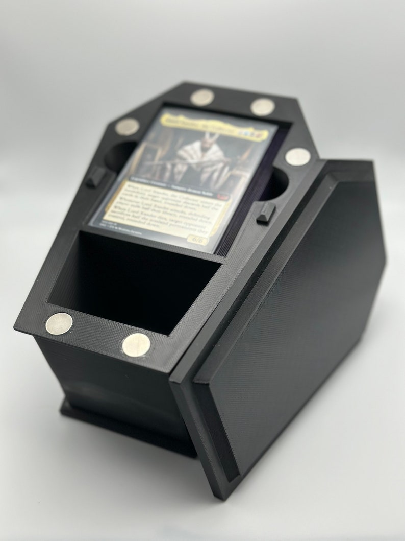 May include: Black coffin-shaped card game deck box with a magnetic closure. The box has a compartment for holding cards and a separate compartment for dice or tokens. The box is made of 3D printed plastic.