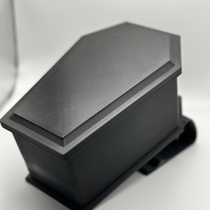 May include: A black coffin-shaped box with a textured surface. The box is closed and has a slightly raised lid.