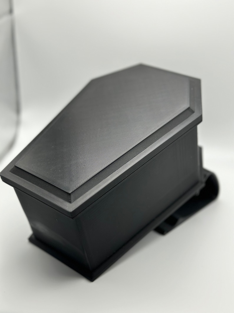 May include: A black coffin-shaped box with a textured surface. The box is closed and has a slightly raised lid.