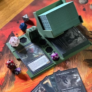 Op de afbeelding: Een groene kaarthouder met meerdere compartimenten, met een grote stapel kaarten. Verschillende veelzijdige dobbelstenen in verschillende kleuren liggen verspreid rond de houder. De kaarten bevatten illustraties en tekst, wat wijst op een verzamelkaartspel.