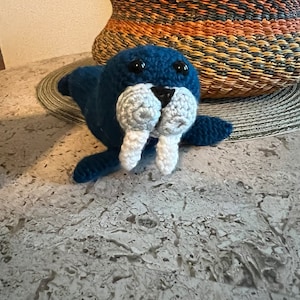 May include: A blue crocheted walrus with white tusks and black eyes. The walrus is lying on a gray textured surface.