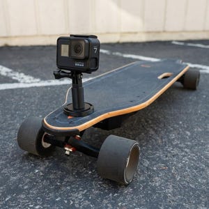 May include: A black electric skateboard with a mounted black GoPro camera. The skateboard has large black wheels and a wooden deck with a black grip tape surface. The camera is mounted on a black stand.