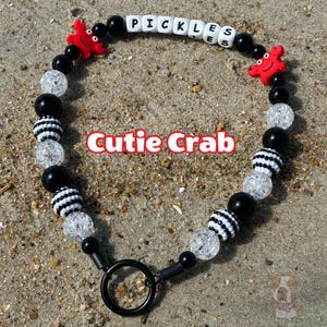 May include: A beaded pet collar with the name "PICKLES" spelled out in white letter beads. The collar features black, white, and clear beads, along with two red crab charms. The text "Cutie Crab" is also visible.