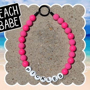 May include: A necklace with bright pink beads and white letter beads spelling "PICKLES". The necklace has a black ring clasp. The image includes a black and white "BEACH BABE" text graphic.