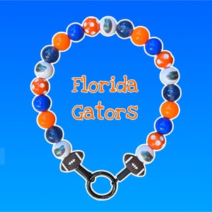 May include: A beaded necklace with blue, orange, and white beads. The necklace has a football-shaped charm on each end and the words "Florida Gators" in orange. The beads are arranged in a circular pattern against a blue gradient background.