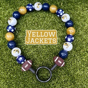 Puede incluir: Un collar de cuentas con temática de fútbol. El collar presenta cuentas azul marino, doradas y blancas alternas con un diseño de chaqueta amarilla. El collar tiene dos dijes en forma de balón de fútbol y un anillo negro. El texto "YELLOW JACKETS" está en el centro.