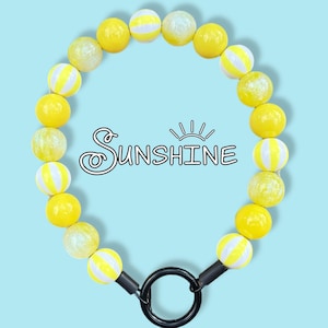 Pode incluir: Um acessório circular de contas alternando entre amarelo sólido, amarelo brilhante e listrado de amarelo e branco. A palavra Sunshine está impressa em preto no centro. Um anel preto está preso na parte inferior com conectores pretos.