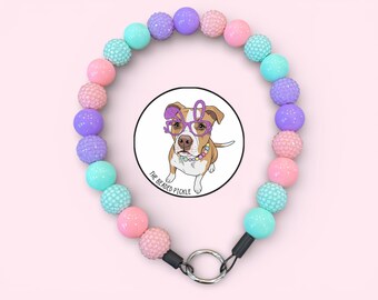 May include: A circular beaded necklace featuring alternating smooth and textured spherical beads in pastel shades of pink, purple, and teal. A central circular pendant displays a brown and white dog wearing purple glasses with the text THE BEADED PICKLE.