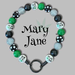 May include: A circular beaded lanyard featuring black, green, silver glitter, and white polka-dot beads. Several white beads are printed with the text I'd Hit That and a green leaf icon. A black metal ring is attached to the bottom of the strand.