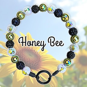 May include: A black and white beaded necklace with a bee charm and a black metal clasp. The beads are black, white, and yellow with a bee design. The necklace is called "Honey Bee".