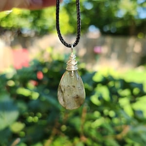 May include: A pendant necklace with a teardrop-shaped, greenish-grey stone. The stone is encased in silver-coloured wire and hangs from a black cord. The background shows blurred green foliage.