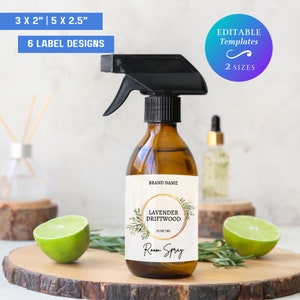 Può includere: Una bottiglia di vetro marrone con un ugello spray nero e un'etichetta bianca. L'etichetta ha un cerchio dorato con il testo "Lavender Driftwood Room Spray" e un design floreale. La bottiglia è appoggiata su una superficie di legno con lime e rosmarino.