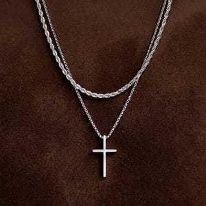 May include: A layered silver-tone necklace set featuring two distinct chains. The top layer is a twisted rope chain, while the bottom layer is a thinner link chain that holds a simple, smooth, vertical silver-tone cross pendant.