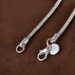 Könnte beinhalten: Eine silberne Schlangenkette mit einem Karabinerverschluss und einem kleinen runden Anhänger, der mit der Zahl 925 geprägt ist. Die Kette hat eine glatte, flexible und abgerundete metallische Textur mit einer polierten Oberfläche.