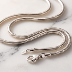 Könnte beinhalten: Eine silberne Schlangenkette mit einem glatten, abgerundeten Glieddesign. Die Kette verfügt über einen Karabinerverschluss und ein kleines rechteckiges Etikett mit der Prägung 925, was auf die Zusammensetzung aus Sterlingsilber hinweist.