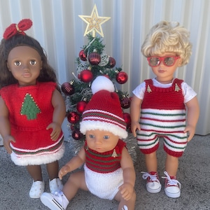 May include: Three dolls dressed in Christmas-themed outfits stand in front of a small Christmas tree. The doll on the left is wearing a red and green dress with a white trim and a gold star on the front. The doll in the middle is wearing a white onesie with red and green stripes and a red and green Santa hat. The doll on the right is wearing a red and green striped outfit with a white trim and sunglasses.