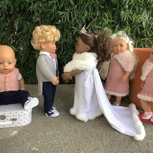 May include: Five dolls dressed in knitted outfits. A baby doll in a pink waistcoat and blue trousers sits on a suitcase. A doll in a grey cardigan and black trousers stands next to a doll in a white dress with a white fur trim. Two dolls in pink dresses with white fur trim stand next to each other.