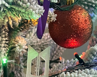 Op de afbeelding: Een transparante glazen kerstbal met een wit, gematteerd, gestileerd logo van de letters "BT" op een paars lint dat aan een besneeuwde kerstboom hangt.
