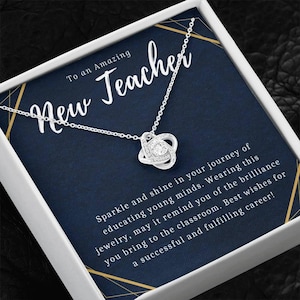 May include: A silver necklace with a heart-shaped pendant, presented in a white box with a navy blue background. The text reads "To an Amazing New Teacher." The card includes a message wishing the recipient a successful career.