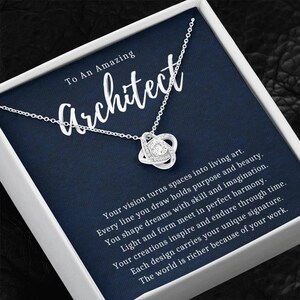 Puede incluir: Un collar plateado con un colgante en forma de nudo, engastado con cristales transparentes. El collar se presenta en una caja blanca con un inserto azul marino. Se lee el texto "To An Amazing Architect".