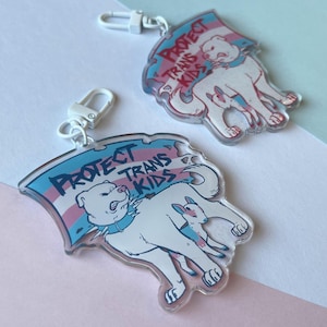 Puede incluir: Dos llaveros de acrílico transparente con un perro blanco y un animal más pequeño, con el texto "PROTECT TRANS KIDS" en una pancarta con los colores de la bandera transgénero. Cada llavero tiene un cierre plateado.