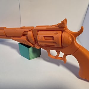 May include: An orange toy gun with a futuristic design. The toy gun is resting on a small teal rectangular block. The gun features a detailed barrel, trigger, and grip, and is made of a solid material.