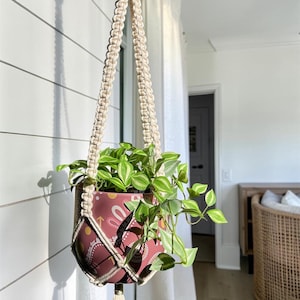 May include: A macrame plant hanger made of off-white cord holding a round, dark pink planter with a white and yellow geometric pattern. A green and white variegated trailing plant grows from the pot, which features a long tassel at the bottom.