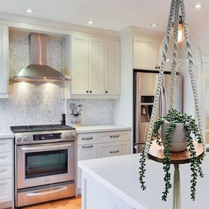 May include: A white macrame plant hanger with a wooden shelf and a small green plant in a grey pot. The hanger is hanging from the ceiling in a kitchen with white cabinets and a stainless steel oven. The text "Little Joyz Studio" is visible in the bottom right corner of the image.
