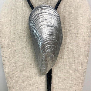 Op de afbeelding: Een bolo-das met een grote, metallic zilverkleurige mosselschelp als centraal hangertje. De schelp vertoont natuurlijke ribbels en een getextureerd oppervlak. Het hangt aan een zwart gevlochten leren koord dat door de bovenkant loopt.