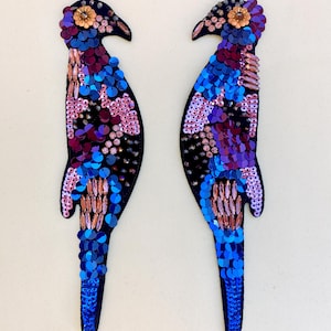May include: Two parrot-shaped appliques with intricate sequin and bead embellishments. The birds feature a colour palette of blue, purple, pink, and orange sequins on a black felt base. The appliques are symmetrical.