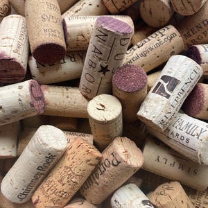 May include: A close-up of a pile of used wine corks, showing a variety of brands and designs. The corks are made of natural cork, with printed text and logos in black ink. The ends of the corks are stained with a reddish-purple hue.