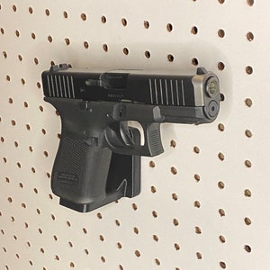 May include: A black handgun with a silver barrel is mounted on a white pegboard. The gun is angled slightly, showcasing its details, including the trigger, grip, and slide. The pegboard has evenly spaced holes.