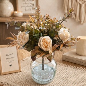 Puede incluir: Un jarrón de cristal transparente lleno de rosas melocotón y vegetación. El jarrón está sobre un mantel individual beige tejido. Un letrero enmarcado dice "HOME sweet HOME". Un arreglo floral decorativo para la decoración del hogar.