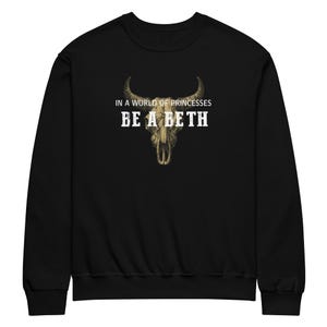 Könnte beinhalten: Schwarzes Sweatshirt mit dem Text "IN A WORLD OF PRINCESSES BE A BETH" über einer detaillierten Illustration eines Kuhschädels. Das Design ist in Weiß und Beige gehalten. Ein lässiges, bequemes Kleidungsstück.