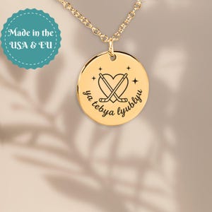 May include: A gold-toned circular pendant necklace featuring an engraved design of a heart with two crossed hockey sticks and four small stars. The text "ya tebya lyublyu" is inscribed in a cursive font along the bottom of the circular charm.