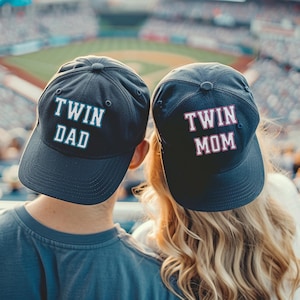 May include: Two navy blue baseball caps with white text that reads "TWIN DAD" and "TWIN MOM". The caps are worn by two people at a baseball game. The background shows the stadium and the crowd.