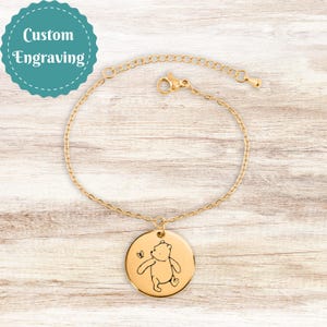 Puede incluir: Pulsera dorada con un colgante redondo con un oso de dibujos animados y una mariposa. La pulsera tiene una cadena delicada y un cierre de mosquetón. La imagen también incluye una insignia turquesa con las palabras "Custom Engraving".