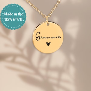 Puede incluir: Collar con colgante dorado con un dije redondo. El dije tiene la palabra "Grammie" en escritura cursiva con un pequeño corazón negro debajo. La cadena también es dorada. Un círculo turquesa con el texto blanco "Made in the USA & EU" es visible.