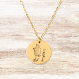 May include: A gold-colored pendant necklace featuring a circular charm with an engraved illustration of a bear and a pig walking together. The necklace has a delicate chain and is set against a light wood grain background.
