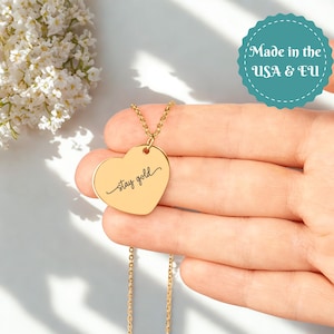 May include: A gold-colored heart-shaped pendant necklace. The pendant has the words "stay gold" in a cursive font. The necklace is on a delicate chain. The background includes white flowers and a teal circular sticker that says "Made in the USA & EU".