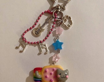 May include: A quirky keychain with a star-shaped clasp, pink beaded chain, and assorted charms. Charms include a guitar, musical notes, and star-shaped beads. The main charm is a cat riding a rainbow-coloured pastry.