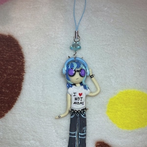 May include: A blue and white figurine of a cartoon character wearing a white t-shirt with the text "I love hot moms" and black pants. The figurine is wearing headphones and has purple eyes. The figurine is attached to a blue and silver phone charm.