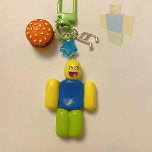 May include: A yellow, blue, and green Roblox character keychain with a burger charm, a star charm, and music note charms. The keychain is attached to a green metal ring.