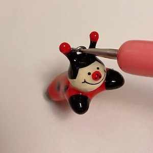 May include: A small ceramic ladybug figurine with black spots and red and white painted details. The ladybug has a silver loop for hanging.