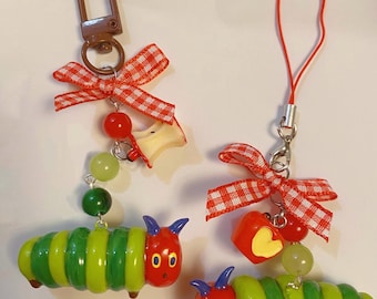 May include: Two green and red caterpillar keychains with red and white gingham bows, red beads, and apple charms. One keychain has a red heart charm and the other has a red apple charm.