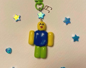 May include: A green and yellow Roblox character keychain with a blue torso and a happy face. The keychain has a green clip and a silver music note charm.