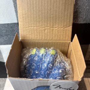 May include: A cardboard box containing blue guitar picks with green tips, wrapped in bubble wrap. The box has a Fender logo on the bottom right corner. The box is open, revealing the contents.