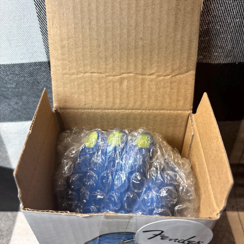 May include: A cardboard box containing blue guitar picks with green tips, wrapped in bubble wrap. The box has a Fender logo on the bottom right corner. The box is open, revealing the contents.